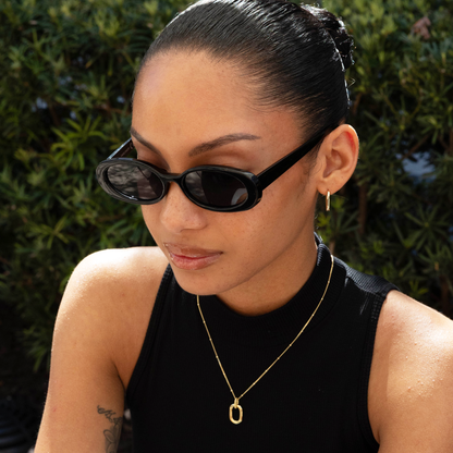 Woman wearing black sunglasses and a black top with a gold oval pendant necklace, standing against a green leafy background.